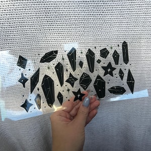 May include: A collection of black crystal and star-shaped decals with white accents. The decals are held against a white knitted fabric background. The crystals vary in size and shape, with a transparent backing.