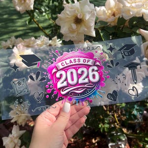 May include: A clear sticker with the text "CLASS OF 2026" in white and pink, surrounded by a splash of blue and pink paint. The sticker also features graduation-themed doodles, including a mortarboard, diploma, and heart.