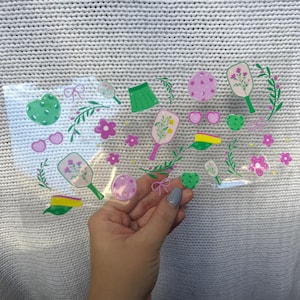 May include: A clear sticker sheet featuring various pastel-colored illustrations. The designs include a green heart, sunglasses, a tennis racket, a skirt, flowers, and a bow. The sticker sheet is held against a white knitted background.