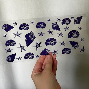 May include: A sheet of dark blue seashell and starfish stickers. The stickers are in various shapes and sizes, including spiral shells and five-pointed stars. A hand is holding one of the stickers.