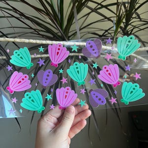 May include: A clear sheet with colorful stickers featuring seashell and butterfly designs in shades of teal, purple, and pink. Small star stickers are also included. The stickers are held by a hand.