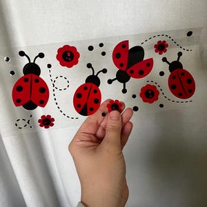 May include: A clear, rectangular sticker sheet featuring red ladybugs with black spots and antennae, along with red flower accents and black dots. The ladybugs are in various poses, some with open wings. A hand holds a small, red flower sticker.