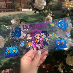 May include: A clear sticker sheet featuring the word "HUNTRAX" in purple and three cartoon girls. The sheet also includes cartoon cats, drinks, and stars in various colors. The sticker is held by a hand.