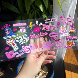 May include: A clear sheet of stickers featuring phrases and images from the movie "Mean Girls." The stickers include quotes like "That's so fetch" and "On Wednesdays we wear pink," along with illustrations of characters and objects.