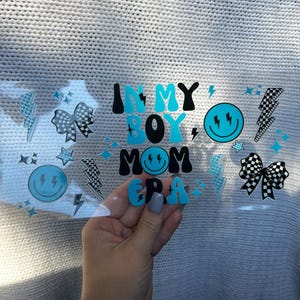 May include: A clear sticker with the text "IN MY BOY MOM ERA" in black and blue letters. The sticker also features lightning bolts, stars, and smiley faces. The sticker is held in a hand.