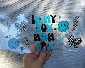 Puede incluir: Una pegatina transparente con el texto "IN MY BOY MOM ERA" en letras negras y azules. La pegatina también presenta rayos, estrellas y caras sonrientes. La pegatina se sostiene en una mano.