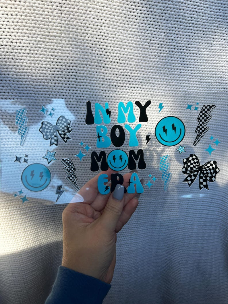 May include: A clear sticker with the text "IN MY BOY MOM ERA" in black and blue letters. The sticker also features lightning bolts, stars, and smiley faces. The sticker is held in a hand.