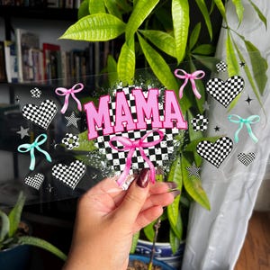 May include: Clear acrylic sign with the word "MAMA" in pink, set against a black and white checkered background. The design includes pink and teal bows, checkered hearts, and stars.