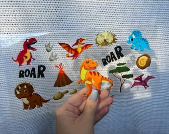 May include: A clear sheet of dinosaur-themed stickers featuring various colorful dinosaurs, footprints, a volcano, and the word "ROAR" in black. The stickers are held in a hand against a white knitted background.