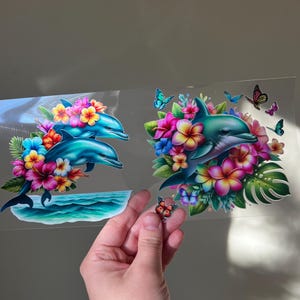 May include: Two dolphin decals with vibrant floral arrangements and butterflies. The dolphins are surrounded by colorful flowers in shades of pink, yellow, and blue. A small butterfly decal is held in a hand.
