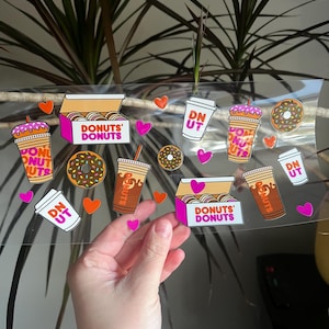May include: A clear sticker sheet featuring colorful illustrations of donuts, coffee cups, and donut boxes. The text "DONUTS' DONUTS" and "DN UT" are visible. Pink hearts and orange hearts are scattered around the images.