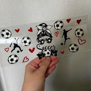 May include: A clear sheet with black and white soccer ball graphics, red hearts, and soccer player silhouettes. The text "Soccer Mom" is also included. The sheet is held by a hand.