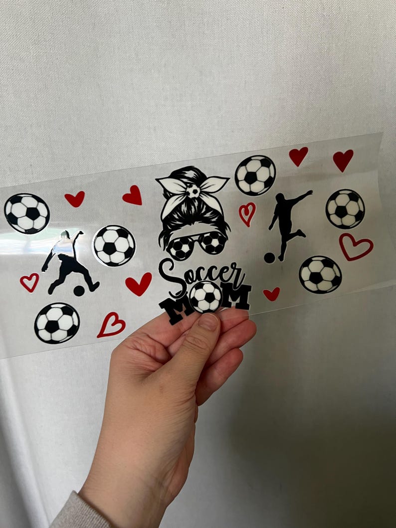 May include: A clear sheet with black and white soccer ball graphics, red hearts, and soccer player silhouettes. The text "Soccer Mom" is also included. The sheet is held by a hand.