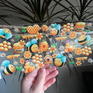 May include: A sheet of clear stickers featuring bee-themed illustrations. The stickers include bees, honey pots, flowers, honeycombs, and a heart-shaped waffle. The word "Honey" is written on a honey pot. The stickers are in shades of yellow, blue, and orange.