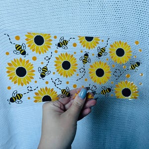 May include: A clear, rectangular sticker sheet featuring a repeating pattern of yellow sunflowers and black and yellow bees. The sunflowers have black centers. The bees have black stripes and wings. The sticker sheet is held against a white knitted background.