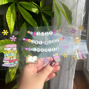 May include: Clear sticker with the text "Just One More Chapter" and illustrations of books and flowers in pink, yellow, and orange. The sticker is held in a hand in front of a green leafy background.