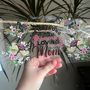 May include: Clear acrylic sign with floral and butterfly accents. The sign features the words "SHE IS STRONG Fearless WARM LOVING PATIENT SELFLESS Mom" in various fonts and colors. Butterflies and flowers are arranged around the edges.