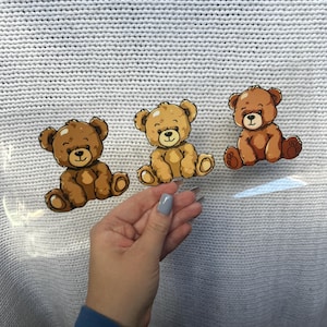 May include: Three cartoon teddy bear stickers in shades of brown and tan are displayed. The bears are sitting with their arms and legs outstretched. The background is a light gray knitted fabric.