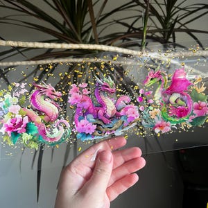 May include: A clear, rectangular sticker featuring three colorful dragons amidst flowers. The dragons are pink, purple, and green, with gold accents. The sticker is held by a hand, showcasing the design's details and vibrant colors.