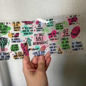 May include: A clear sticker sheet featuring colorful Bible affirmations. The text includes phrases like "God is Love" and "The Lord is my Shepherd." The design incorporates floral and cross illustrations, with a pink and green color scheme.