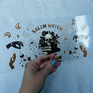 May include: A clear sticker with a gold and black design featuring a skull, roses, hands, moons, and the words "Salem Witch Book Club." The sticker is held in a hand against a white knitted background.