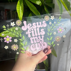 May include: A clear sticker with the phrase "Jesus fills my cup" in pink bubble letters, surrounded by floral and cross graphics in pastel colors. The sticker is held up against a background of green leaves.