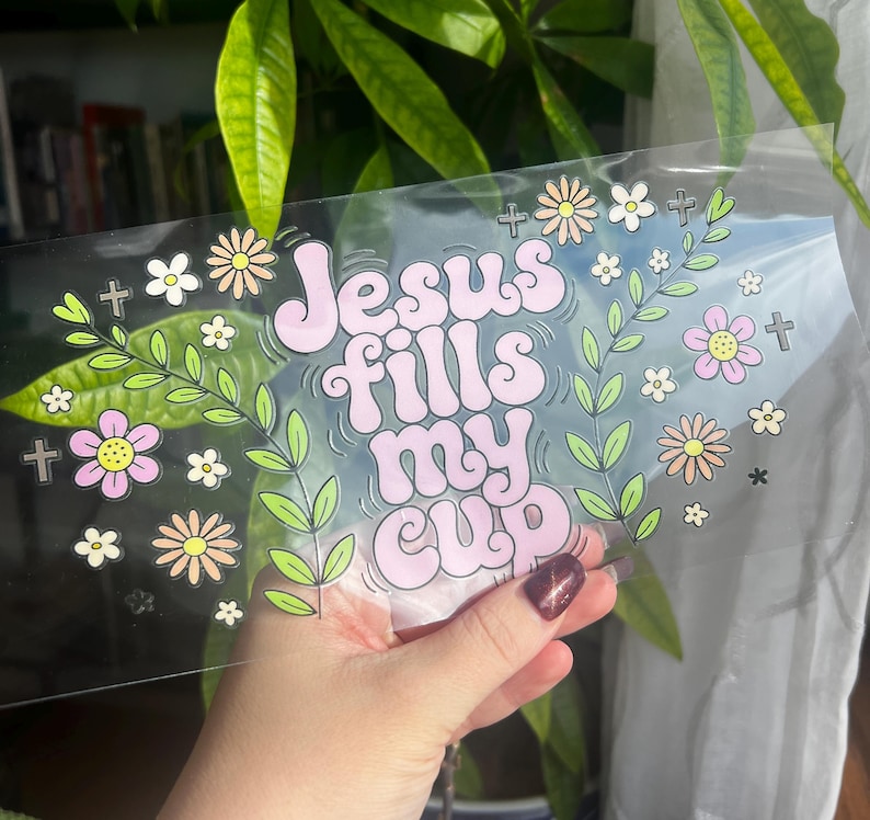 May include: A clear sticker with the phrase "Jesus fills my cup" in pink bubble letters, surrounded by floral and cross graphics in pastel colors. The sticker is held up against a background of green leaves.