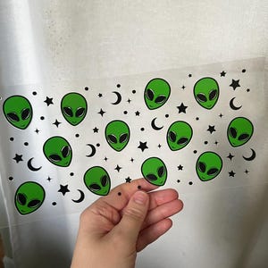 May include: A clear sticker sheet featuring a repeating pattern of green alien faces, black stars, and crescent moons. The alien heads have large black eyes and are set against a white background. The sheet is held by a hand.