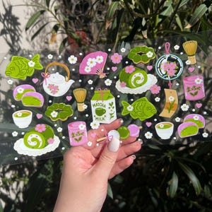 May include: A clear sticker sheet featuring various Japanese-themed illustrations. Designs include matcha tea, fish-shaped cakes, a fan, a teapot, and a milk carton. The color palette consists of green, pink, white, and brown accents. Small white flower details are scattered throughout.