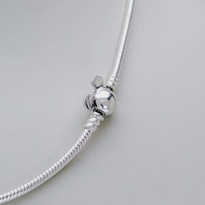 May include: A silver-colored necklace with a Mickey Mouse-shaped clasp. The clasp is a round bead with two small, round ears on top. The necklace has a snake chain design.
