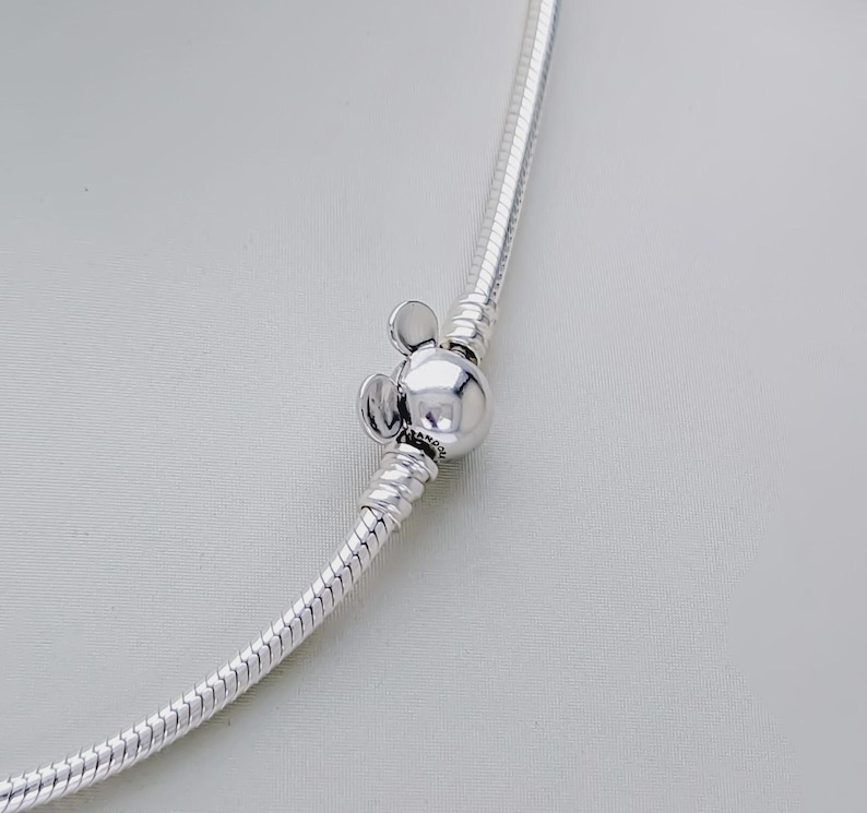 May include: A silver-colored necklace with a Mickey Mouse-shaped clasp. The clasp is a round bead with two small, round ears on top. The necklace has a snake chain design.