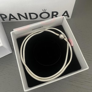 May include: A silver snake chain bracelet in a white box with a black interior. The bracelet is coiled within the box, which sits on a grey surface. A white shopping bag with the brand name "Pandora" is visible in the background.