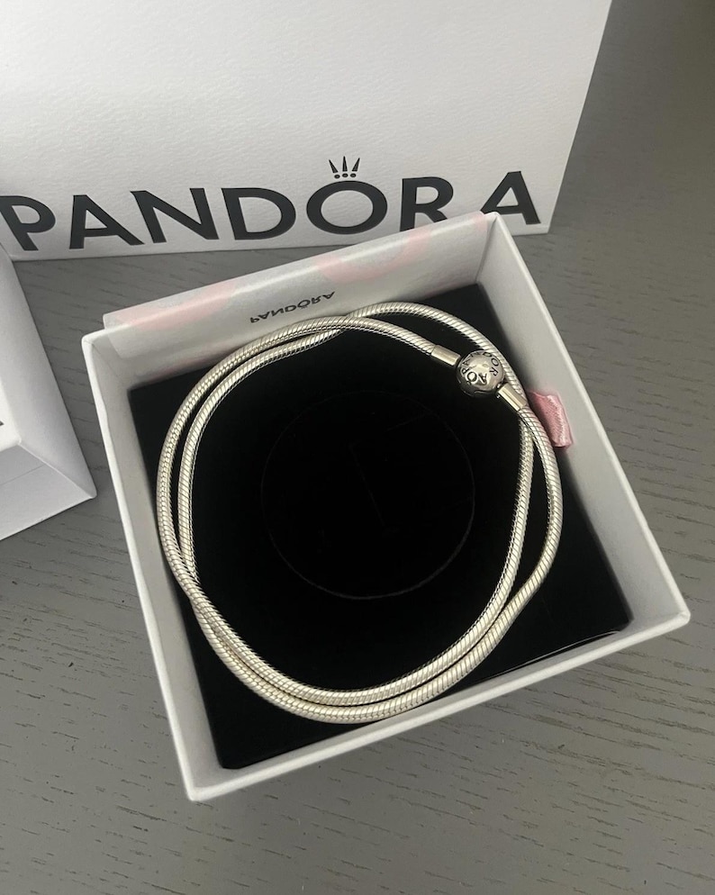 May include: A silver snake chain bracelet in a white box with a black interior. The bracelet is coiled within the box, which sits on a grey surface. A white shopping bag with the brand name "Pandora" is visible in the background.