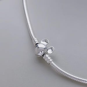 May include: A silver necklace with a Mickey Mouse charm. The charm is a round bead with two small, round ears. The necklace has a snake chain design. The Disney logo is visible on the charm.