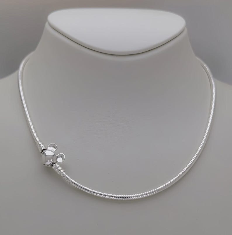 May include: A silver necklace with a Mickey Mouse-shaped charm. The necklace is displayed on a white mannequin. The charm is a detailed, three-dimensional design. The necklace has a snake chain style.