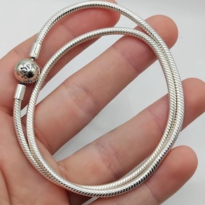 May include: A silver snake chain bracelet with a round charm. The bracelet is made of a flexible, ribbed metal and is held in a hand. The charm has the word "Pandora" engraved on it.