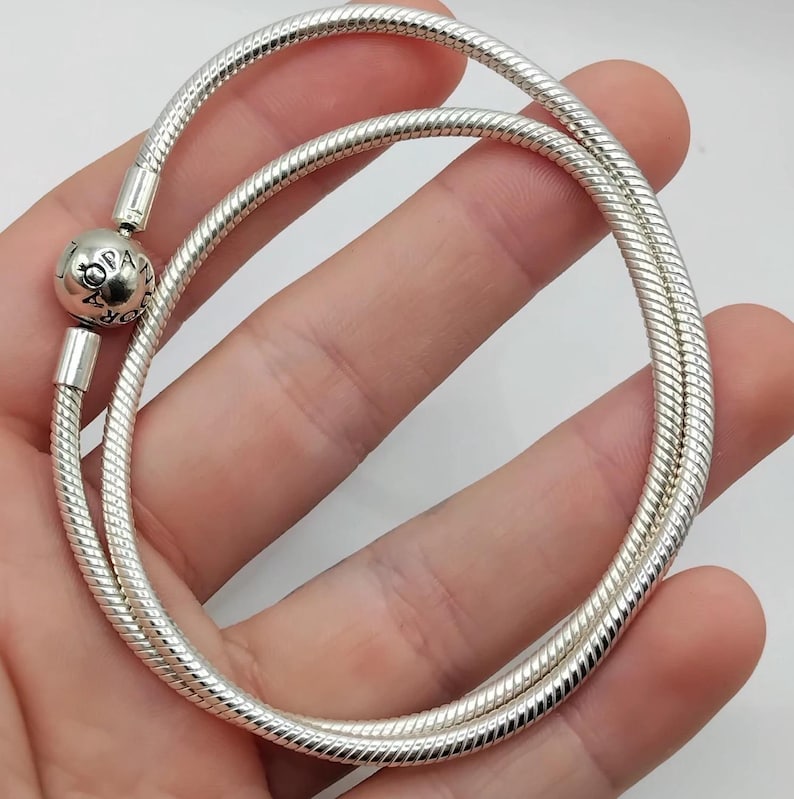 May include: A silver snake chain bracelet with a round charm. The bracelet is made of a flexible, ribbed metal and is held in a hand. The charm has the word "Pandora" engraved on it.