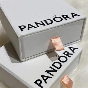 May include: Two white Pandora boxes stacked on top of each other. The boxes have black text that reads "PANDORA" and a small crown logo. Each box has a peach-colored ribbon pull.