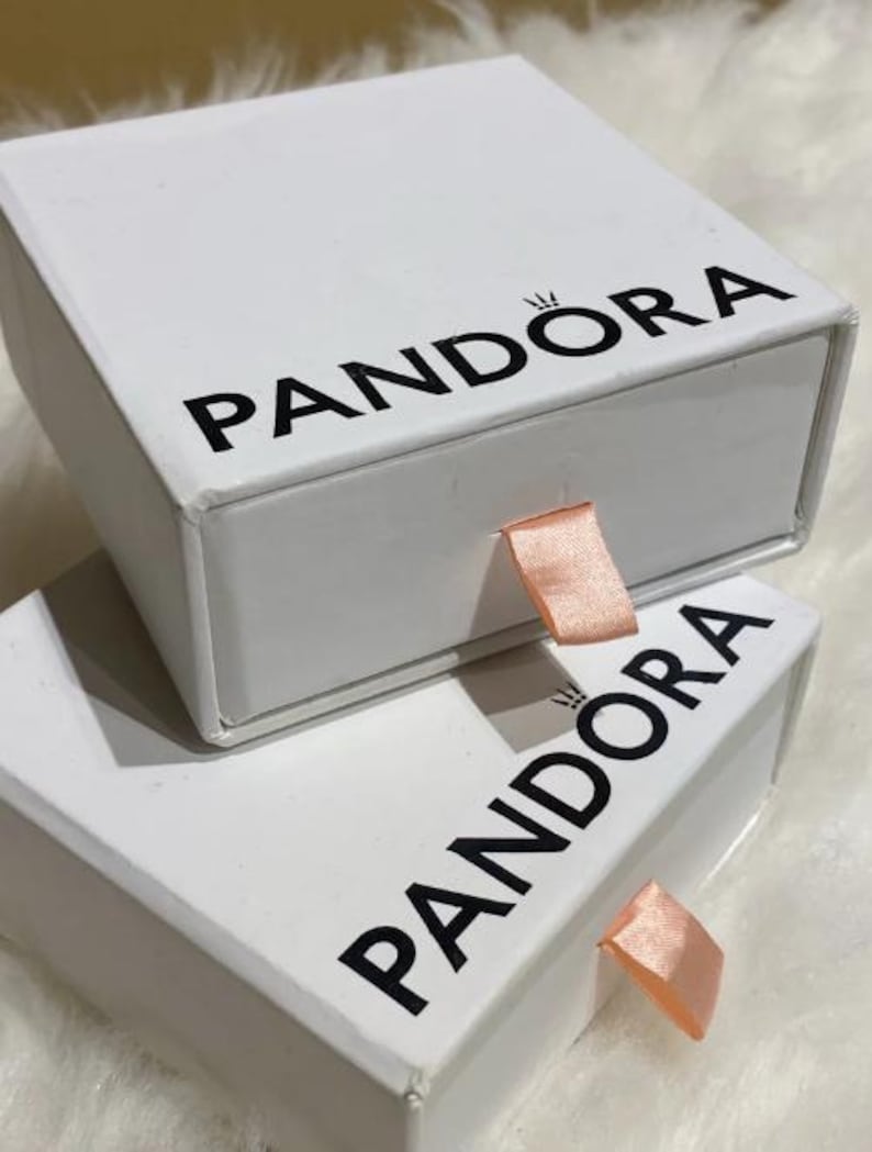 May include: Two white Pandora boxes stacked on top of each other. The boxes have black text that reads "PANDORA" and a small crown logo. Each box has a peach-colored ribbon pull.