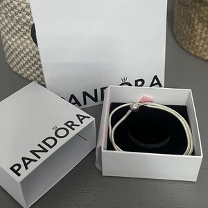 May include: A silver bracelet with a round charm is presented in an open white box with a black interior. The box is inside a larger white box with the brand name "PANDORA" printed on it. A woven basket is in the background.