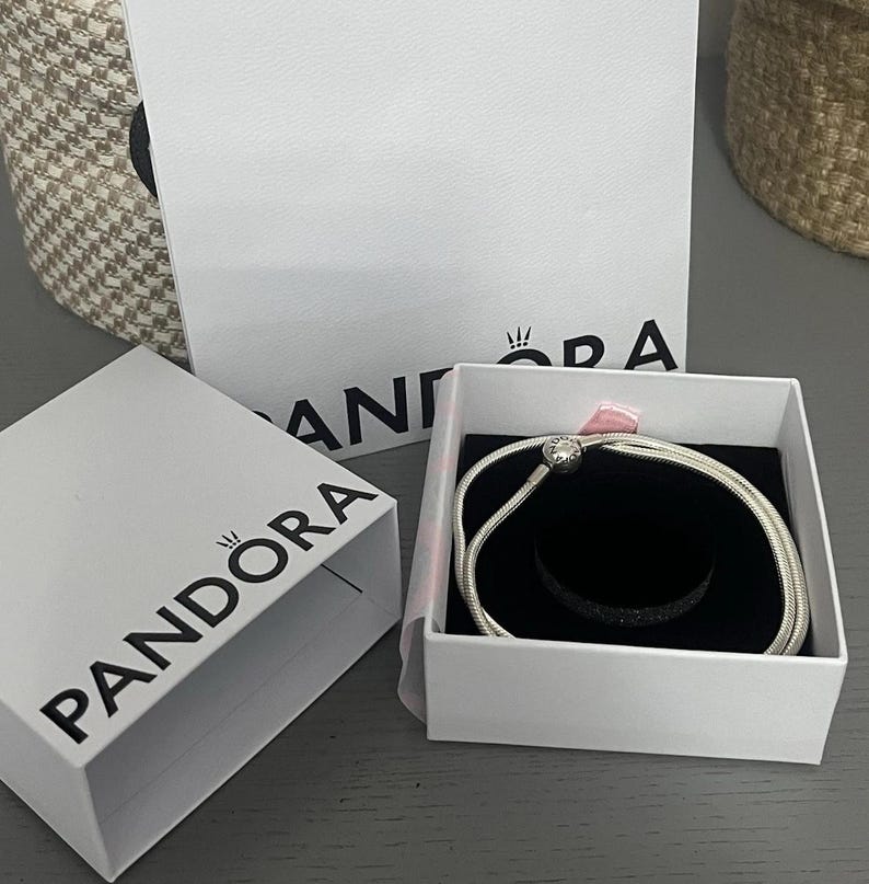 May include: A silver bracelet with a round charm is presented in an open white box with a black interior. The box is inside a larger white box with the brand name "PANDORA" printed on it. A woven basket is in the background.