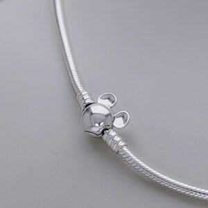 May include: A silver necklace with a Mickey Mouse charm. The charm is a round bead with two small, round ears. The necklace has a snake chain design. The Disney logo is visible on the charm.