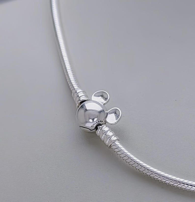 May include: A silver necklace with a Mickey Mouse charm. The charm is a round bead with two small, round ears. The necklace has a snake chain design. The Disney logo is visible on the charm.