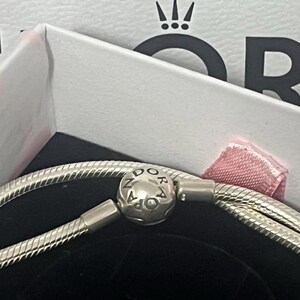 May include: A silver bracelet with a round clasp bearing the brand name "PANDORA". The bracelet is presented in a white box with a pink ribbon and the brand's logo. The bracelet has a snake chain design.