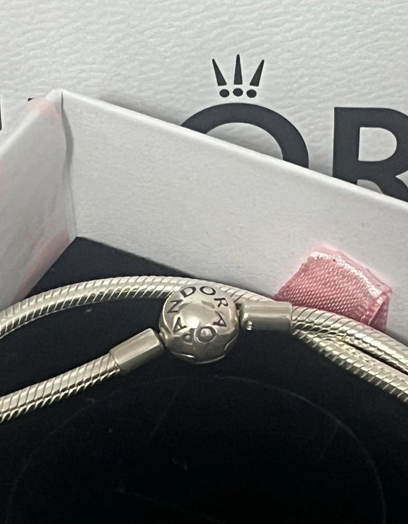 May include: A silver bracelet with a round clasp bearing the brand name "PANDORA". The bracelet is presented in a white box with a pink ribbon and the brand's logo. The bracelet has a snake chain design.