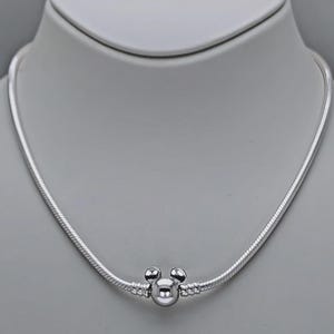May include: A silver necklace with a Mickey Mouse charm. The necklace has a snake chain design and a round charm with two small spheres on top, resembling Mickey Mouse's ears. The necklace is displayed on a white mannequin.