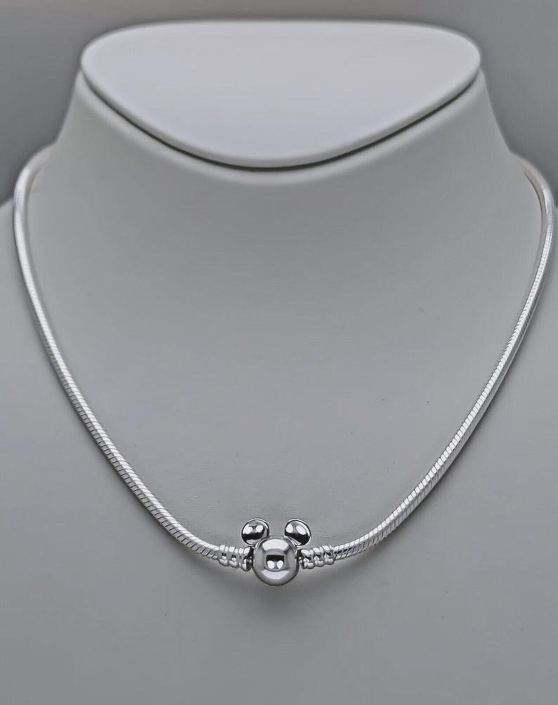May include: A silver necklace with a Mickey Mouse charm. The necklace has a snake chain design and a round charm with two small spheres on top, resembling Mickey Mouse's ears. The necklace is displayed on a white mannequin.