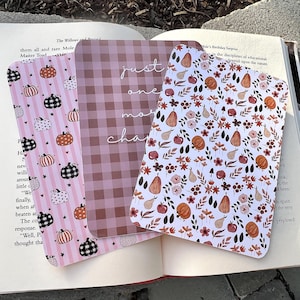 May include: Three rectangular bookmarks resting on an open book. One features pink stripes and black and orange pumpkins. The center bookmark has a brown gingham pattern with the text just one more chapter. The third has a floral and pumpkin print.