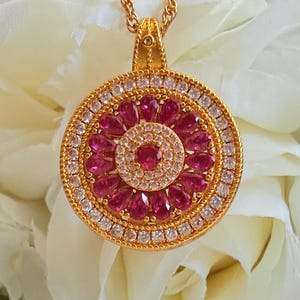 May include: A gold-toned pendant necklace with a circular design. The pendant features a central cluster of clear stones surrounded by a ring of pink gemstones and an outer ring of clear stones. The necklace chain is also gold-toned.