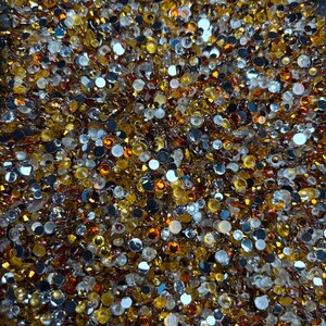 May include: A close-up of a collection of small, round rhinestones. The rhinestones are a mix of gold, silver, and amber colours. They are scattered closely together, creating a sparkling, textured surface. Suitable for craft projects.